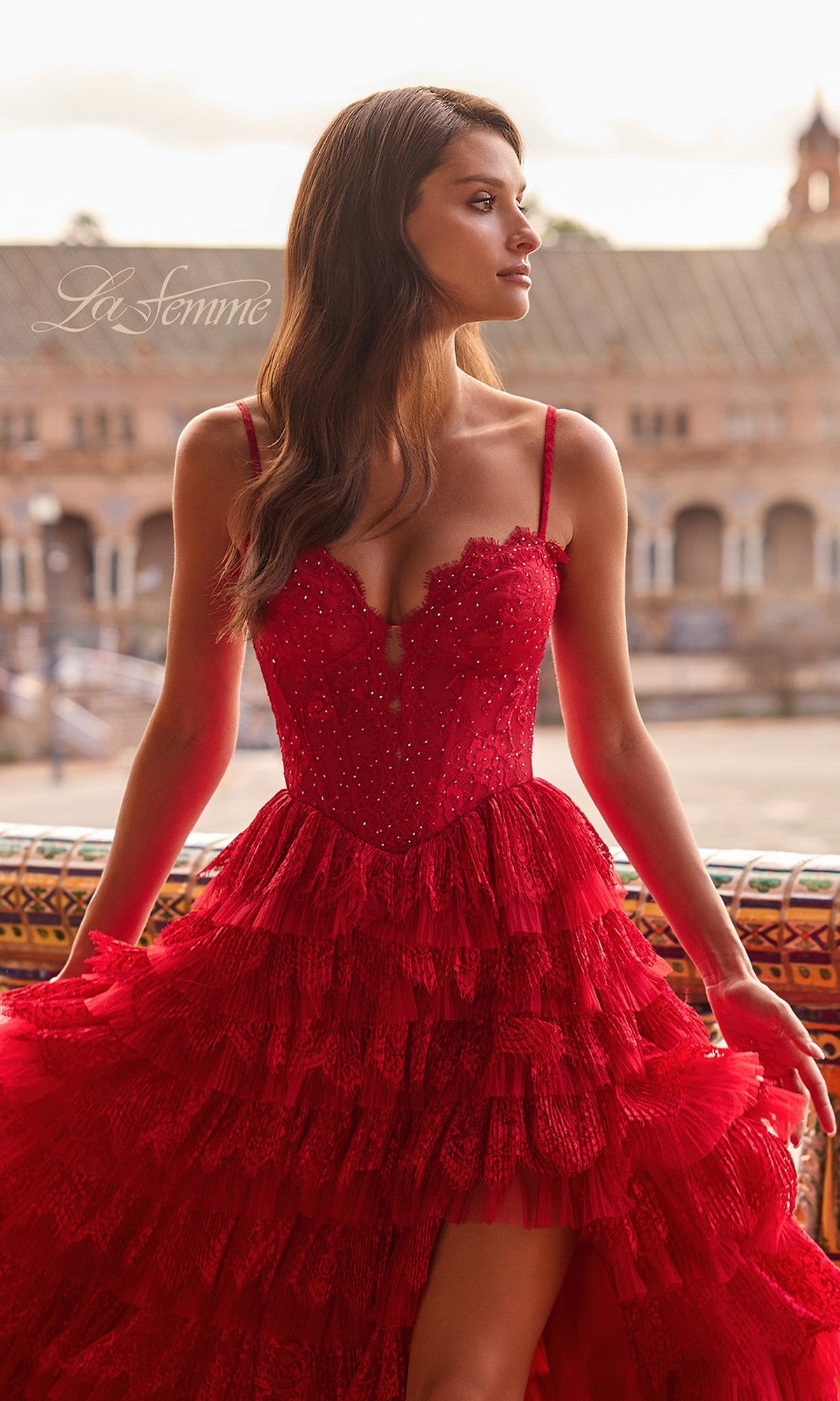 A woman wearing the La Femme 33525 long prom dress with ruffled spaghetti straps in bright red stands outdoors, looking to the side, with a historic building softly blurred in the background.